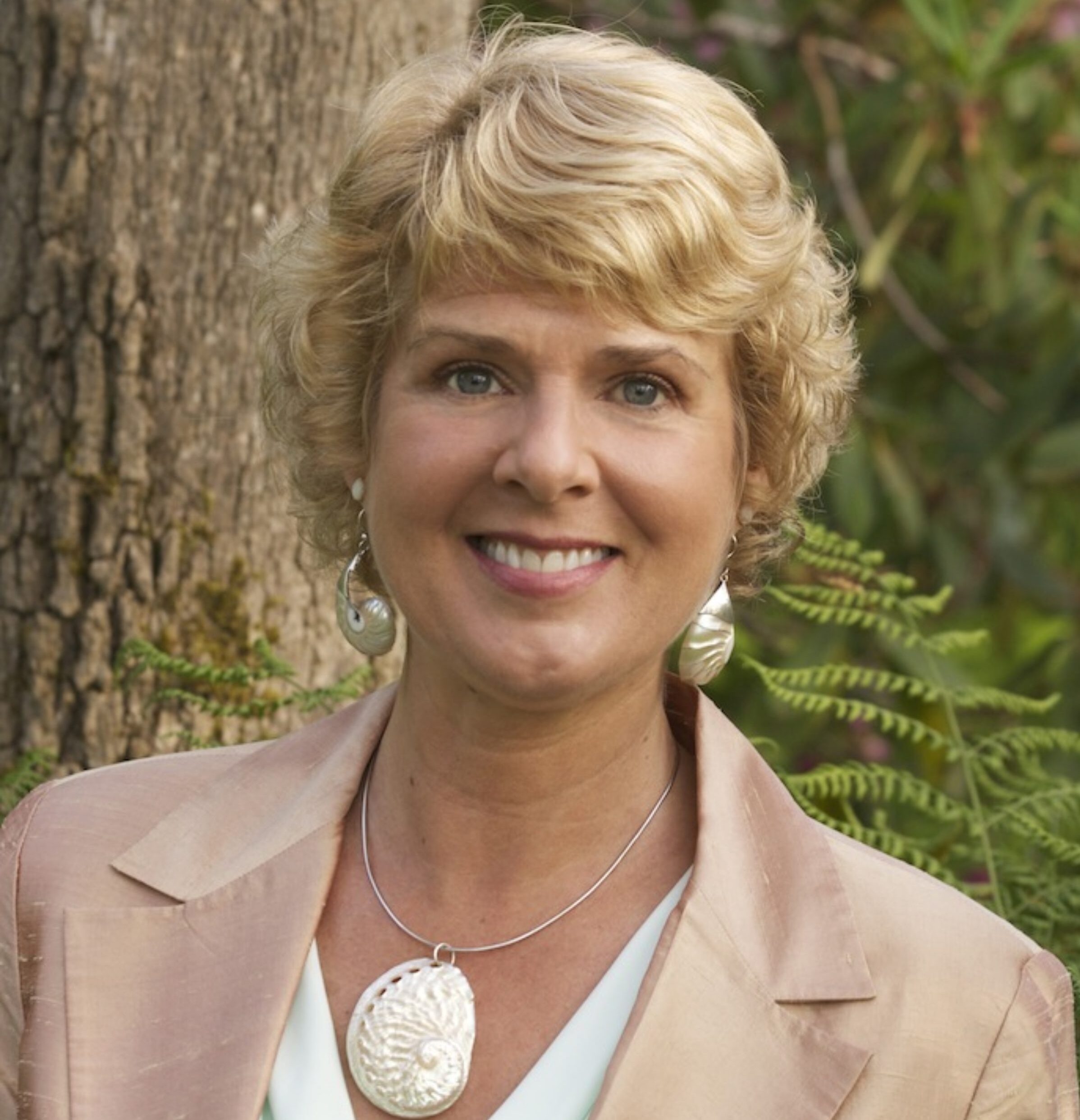 A woman in a beige jacket standing next to a tree.